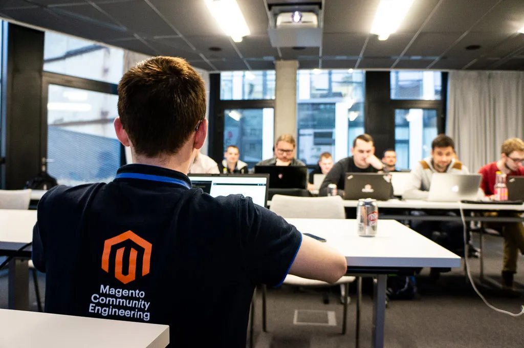 Classroom, in the foreground a teacher sitting rear-facing in T-shirt with Magento logo and Magento Community Engineering text on it, in the background - people in front of their laptops sitting at the tables in two rows.