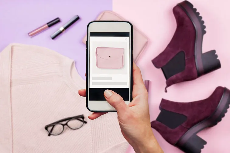 A hand holding a smartphone with a picture of a pink bag on the screen. In the background - clothes and accessories: shoes, lipstick, bag (the same as on the phone screen), sweater and glasses. Everything in rose-purple colors.