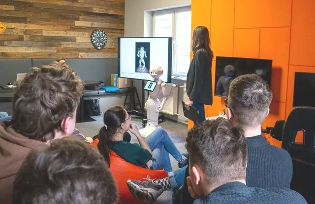 Group of people seated and watching a presentation. In the background a girl with robot Pepper and presentation screen.