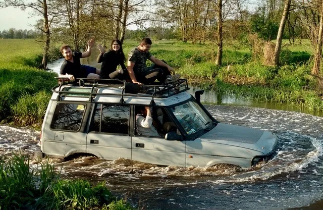 Three people in outdoor clothing sitting on the roof of an off-road vehicle crossing a river.