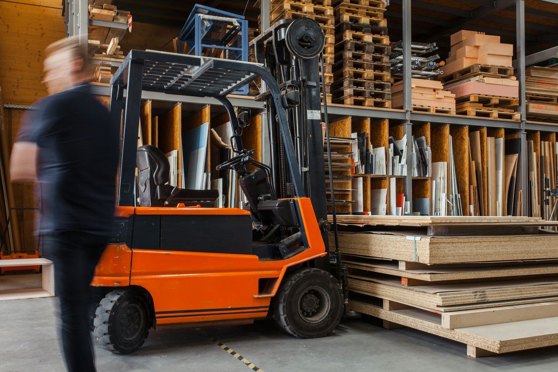 man walking by a forklift in front of shelves stacked with boxes and other containers of inventory